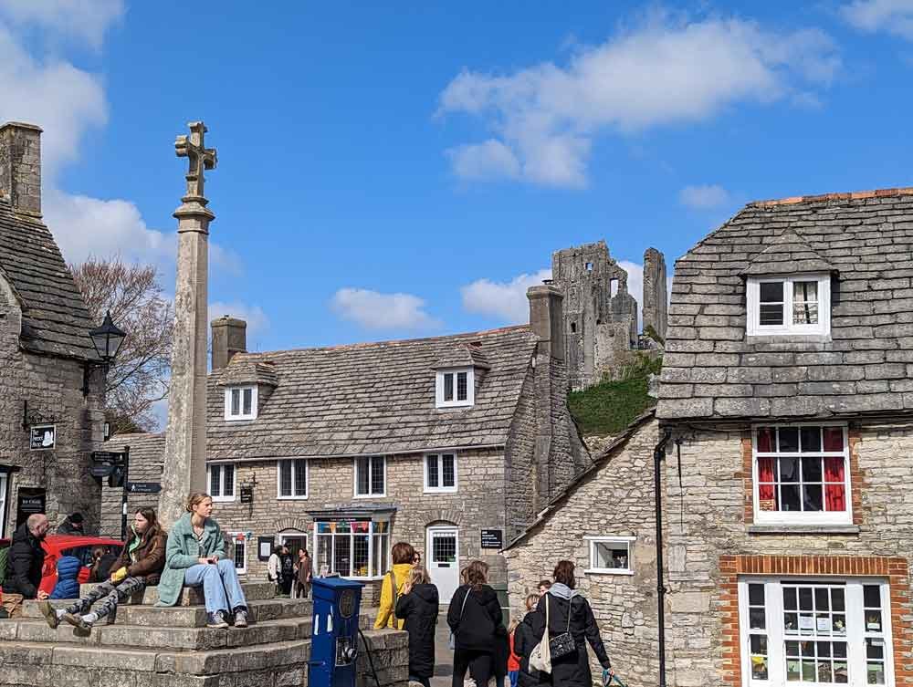 Corfe Castle Village, People sitting on stone cross memorial in front of stone homes overlooked by Corfe Castle.