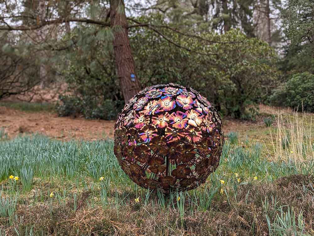 Bronze sphere shaped sculpture in gardens with trees behind.