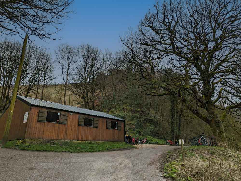 Brown wooden hut with trees in backround and gravel road in front. This is the Blackwell Mill Cycle Hire, Monsal Trail, Peak District