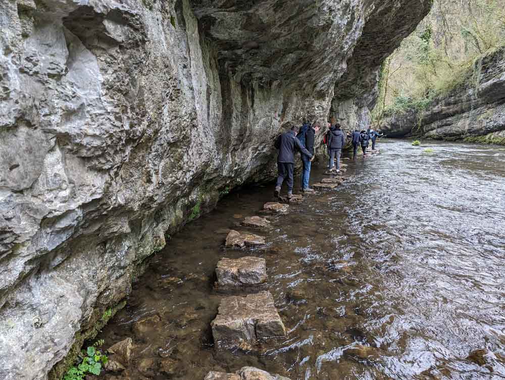 Backs of people crossing the stepping stones under overhanging limestone cliff.