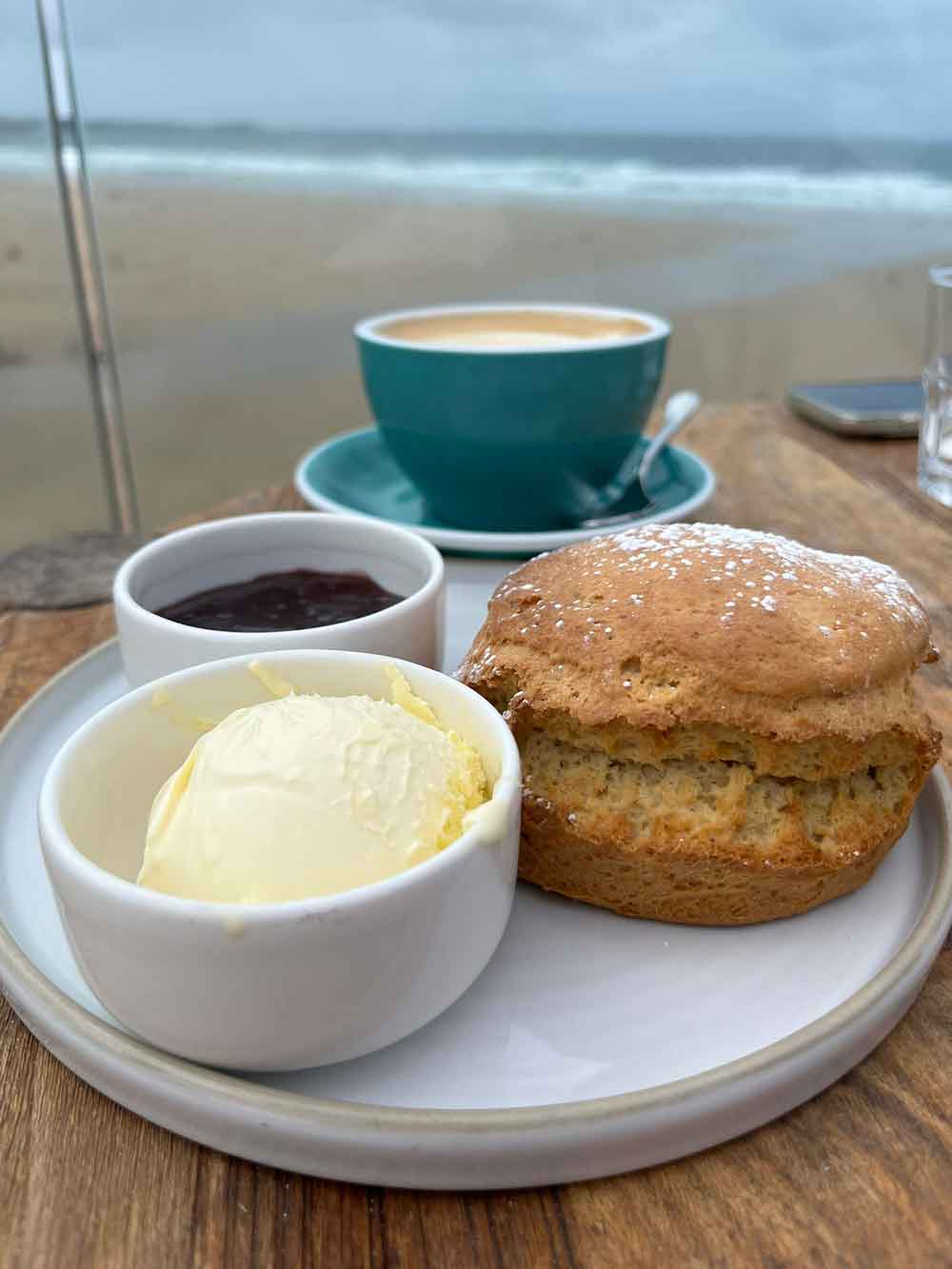 Cream tea (scones, cream and jam) in front of coffee cup looking out over Watergate Bay