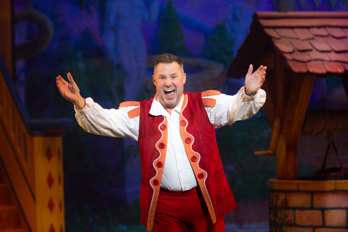 Aaron James as Muddles in Snow White, wearing red velvet trousers, a matching waistcoat and a long white shirt, stood next to a well