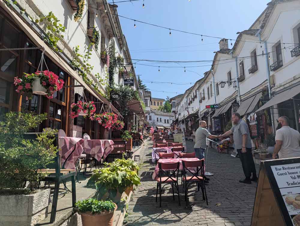 Historic streets with restaurant tables and chairs in centre, 2 men greeting and flower pots
