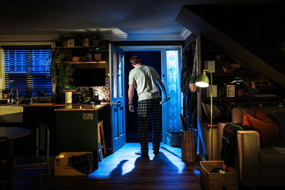Man holding baseball bat in dark looking out of house into garden.