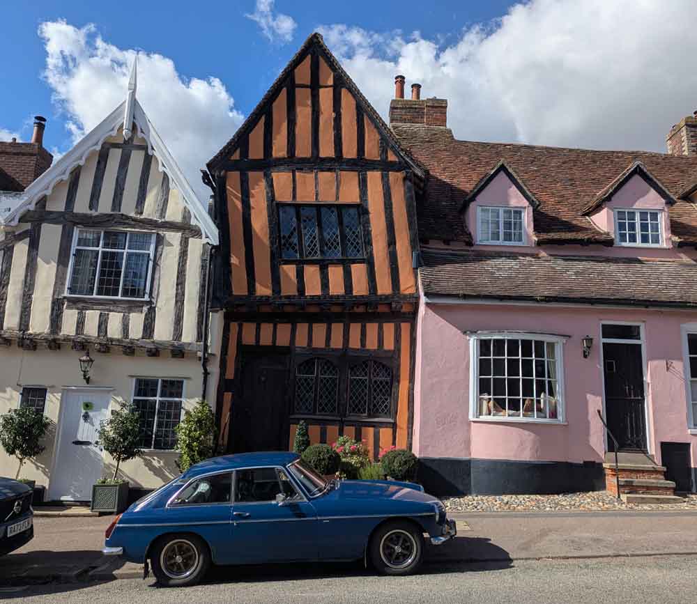 The orange timber-framed Crooked House in Lavenham with a pink house and white house on either side and a blue MG in foreground